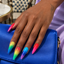 Rainbow glitter stiletto nails worn on a hand resting on a cobalt handbag